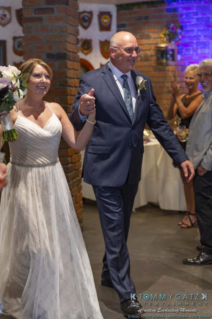 Grace-Michael-063018-RECEPTION-Low-Res-WM-278 - TOMMY GATZ ENTERTAINMENT A bride and groom holding hands enter a reception venue, smiling. The bride holds a bouquet, and guests are visible in the background.