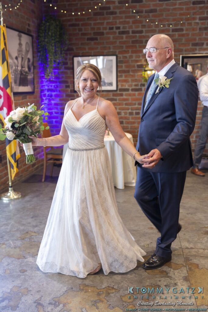 Grace-Michael-063018-RECEPTION-Low-Res-WM-284 - TOMMY GATZ ENTERTAINMENT A couple smiling and holding hands, dressed in wedding attire. She holds a bouquet, and decorations are visible in the background.