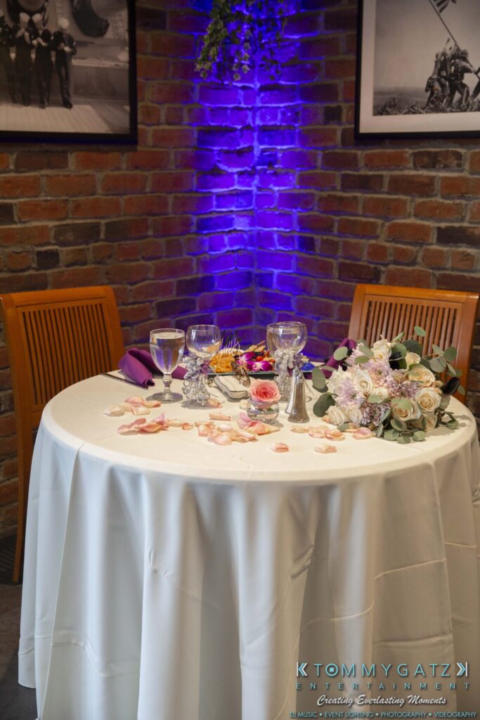 Grace-Michael-063018-RECEPTION-Low-Res-WM-299 - TOMMY GATZ ENTERTAINMENT A round table set for a romantic meal with two chairs, featuring a white tablecloth, floral centerpiece, wine glasses, and scattered rose petals. A brick wall and framed photos are in the background.