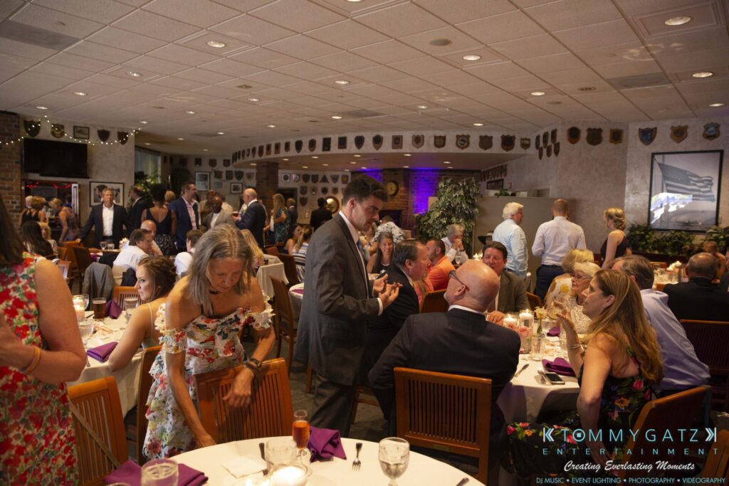 Grace-Michael-063018-RECEPTION-Low-Res-WM-308 - TOMMY GATZ ENTERTAINMENT A group of people socializing and dining at a formal indoor event. Tables are set with candles and purple napkins. A banner reading "Tommy Gatz Entertainment" is visible.