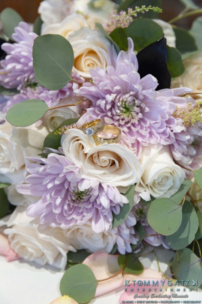 A bouquet of purple chrysanthemums, pale roses, and greenery with two gold rings placed on top.