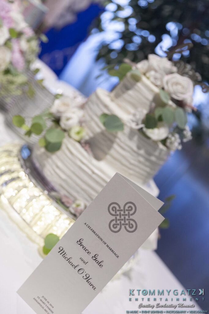 Wedding reception with a tiered cake decorated with flowers and greenery. A program reads "A Celebration of Marriage, Grace Lake and Michael O'Hara.