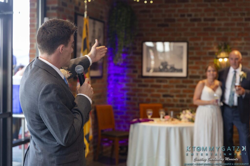 Grace-Michael-063018-RECEPTION-Low-Res-WM-365 - TOMMY GATZ ENTERTAINMENT A man in a suit speaks with a microphone in a brick-walled room. A bride and groom stand at a table in the background, listening.