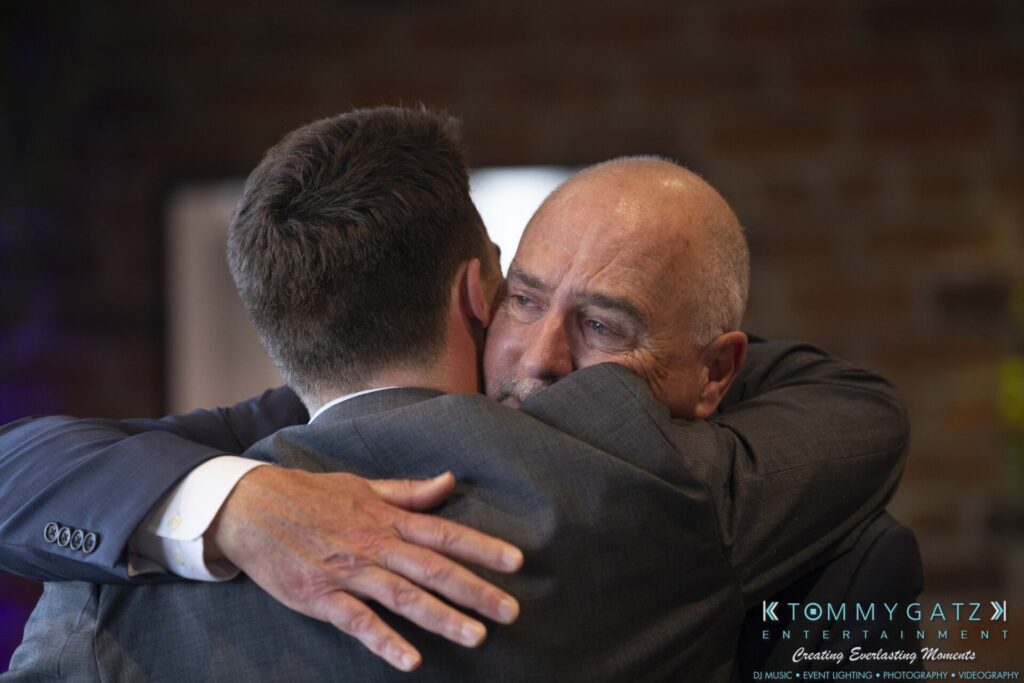 Grace-Michael-063018-RECEPTION-Low-Res-WM-374 - TOMMY GATZ ENTERTAINMENT Two men hugging in an emotional embrace, wearing suits, in a softly lit setting with a blurred background.