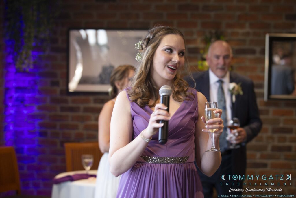 Grace-Michael-063018-RECEPTION-Low-Res-WM-377 - TOMMY GATZ ENTERTAINMENT A woman in a purple dress holds a microphone and a glass, speaking at an event with people in the background.