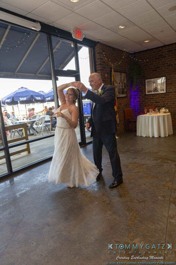 Grace-Michael-063018-RECEPTION-Low-Res-WM-386 - TOMMY GATZ ENTERTAINMENT A couple dances together in a room with large windows and a brick wall. The woman wears a white dress, and the man is in a suit. A decorated table is visible in the background.