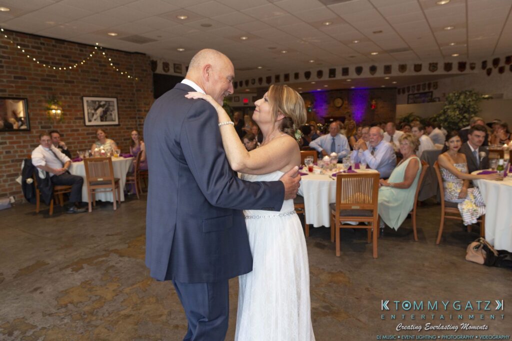 Grace-Michael-063018-RECEPTION-Low-Res-WM-396 - TOMMY GATZ ENTERTAINMENT A bride and groom share a first dance in a warmly lit reception room, surrounded by seated guests and decorative string lights.