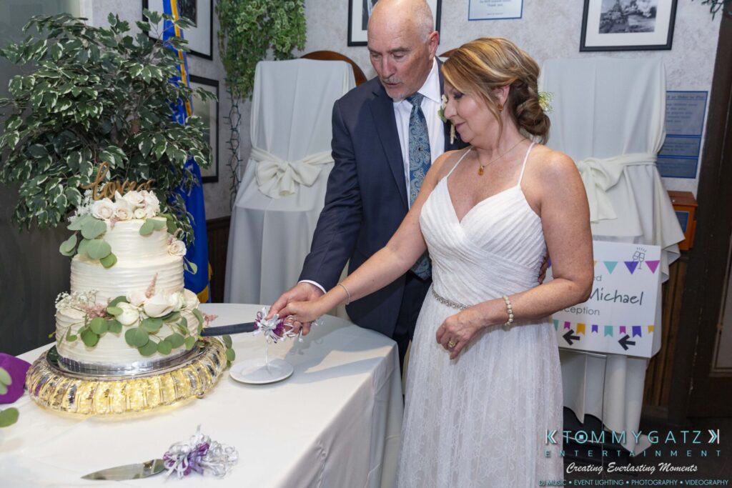 Grace-Michael-063018-RECEPTION-Low-Res-WM-410 - TOMMY GATZ ENTERTAINMENT A couple in formal attire cuts a three-tiered white cake decorated with greenery and flowers at an indoor event.