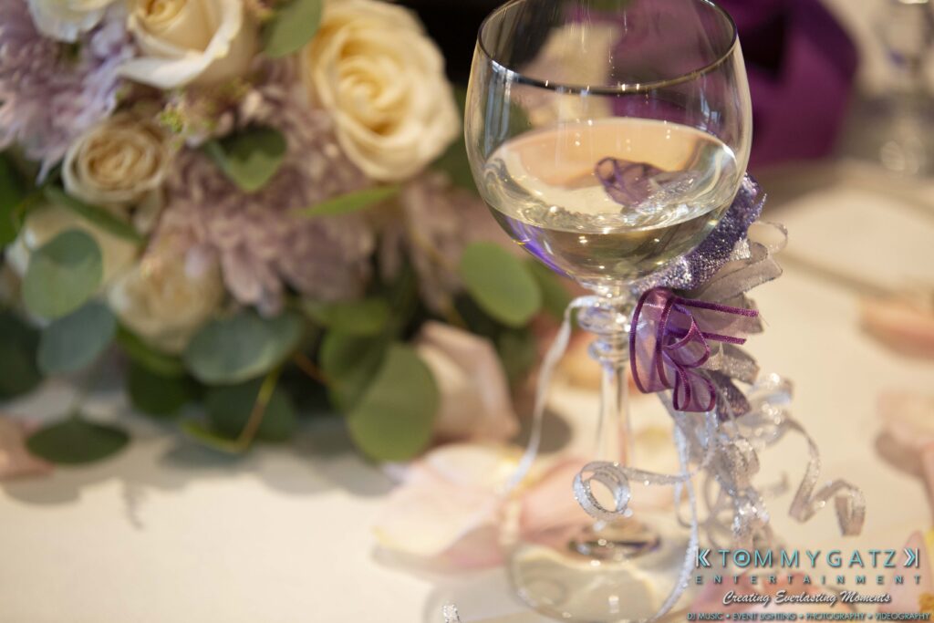A wine glass with decorative ribbons sits on a table, accompanied by a floral arrangement of roses and purple flowers.