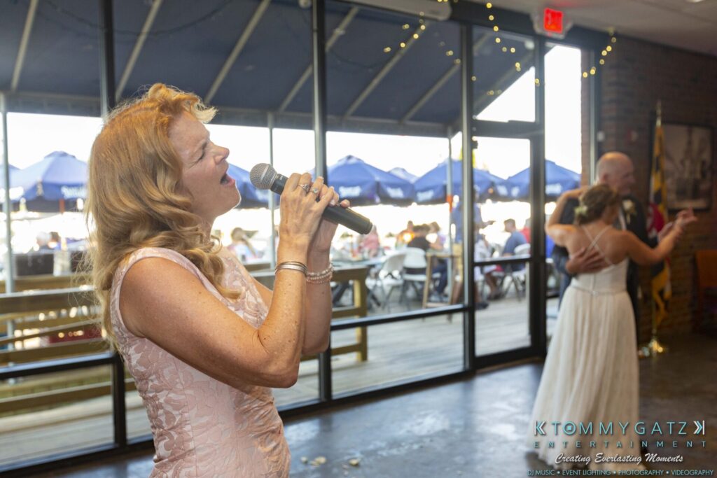 Grace-Michael-063018-RECEPTION-Low-Res-WM-443 - TOMMY GATZ ENTERTAINMENT A woman sings into a microphone at an indoor event, while a couple dances in the background near large windows with outdoor seating visible.