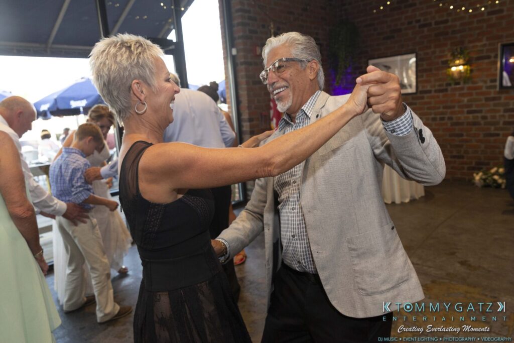 Grace-Michael-063018-RECEPTION-Low-Res-WM-457 - TOMMY GATZ ENTERTAINMENT Two people dancing joyfully in a decorated venue with other people in the background.