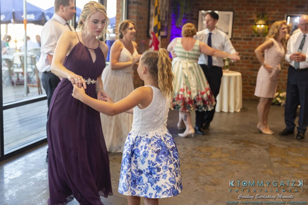 Grace-Michael-063018-RECEPTION-Low-Res-WM-474 - TOMMY GATZ ENTERTAINMENT A woman and a young girl dance together at an indoor event with people socializing in the background.