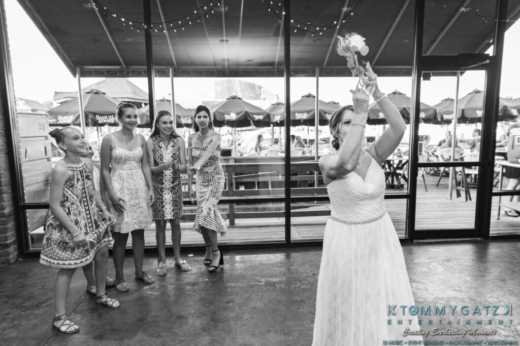 Grace-Michael-063018-RECEPTION-Low-Res-WM-482 - TOMMY GATZ ENTERTAINMENT A bride prepares to throw her bouquet in a room with a glass wall. Five women stand in the background, ready to catch it.