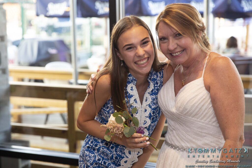 Grace-Michael-063018-RECEPTION-Low-Res-WM-485 - TOMMY GATZ ENTERTAINMENT A young woman in a blue dress and an older woman in a white dress smile while posing together indoors. The younger woman holds a small bouquet.