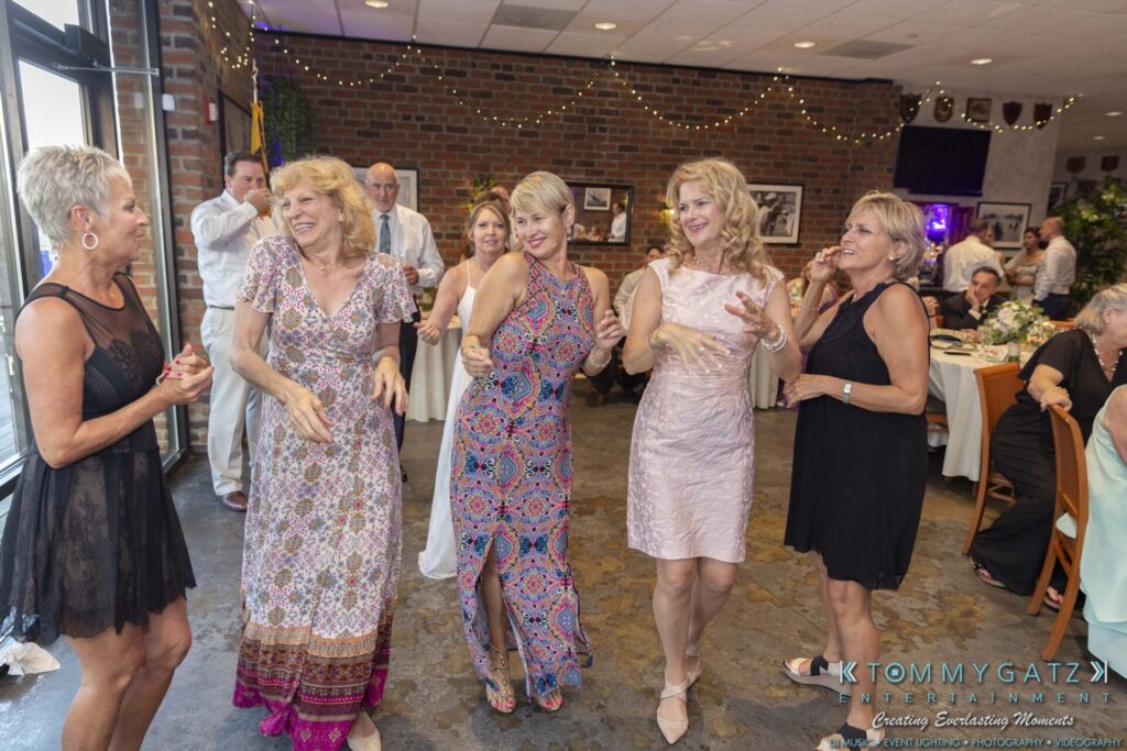 Grace-Michael-063018-RECEPTION-Low-Res-WM-491 - TOMMY GATZ ENTERTAINMENT A group of women in dresses dancing energetically at an indoor event, with tables and guests in the background.
