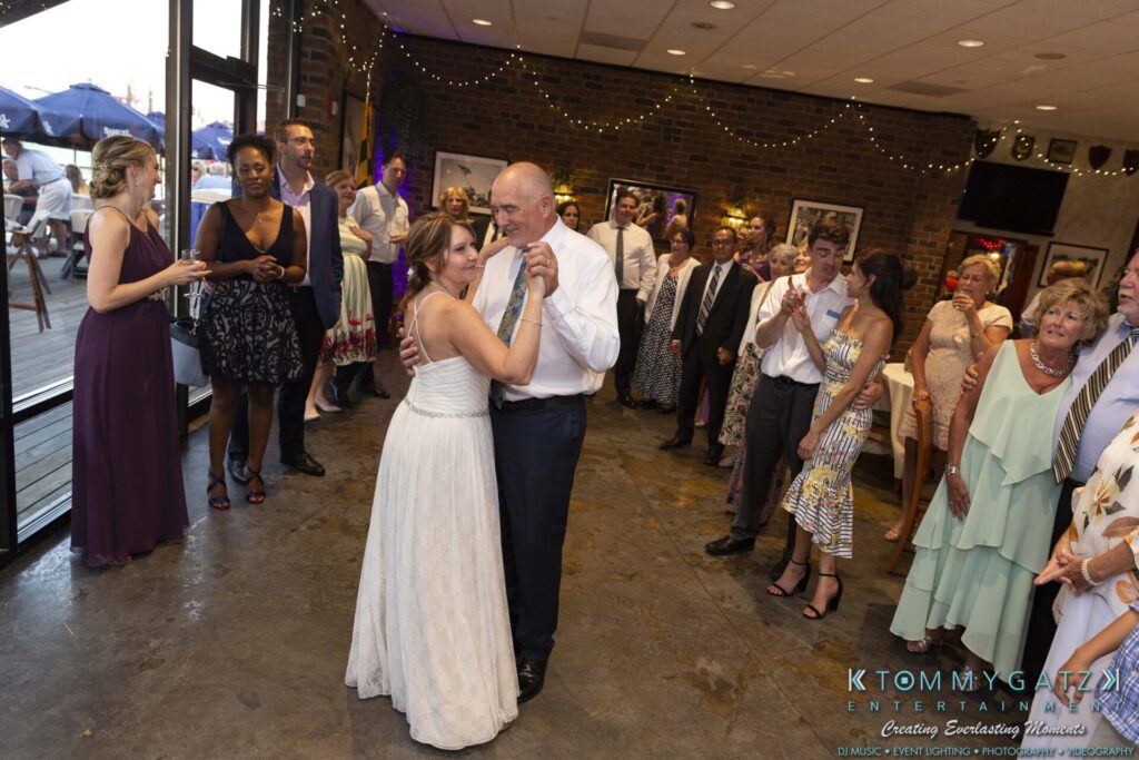 A couple dances in the center of a reception hall, surrounded by guests. The room is decorated with string lights and framed photos.