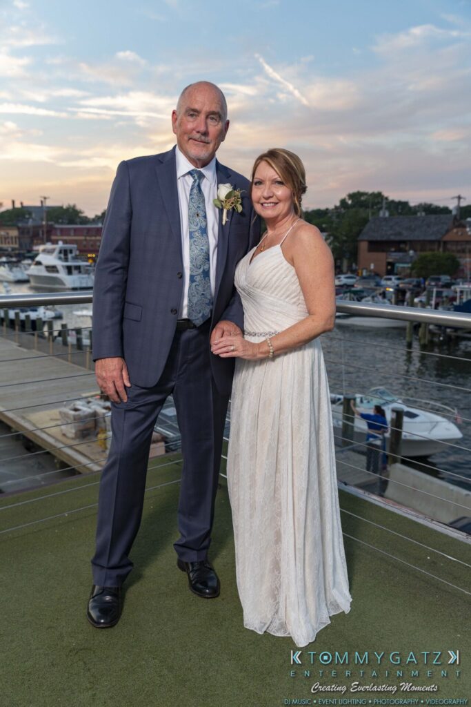 Grace-Michael-063018-RECEPTION-Low-Res-WM-506 - TOMMY GATZ ENTERTAINMENT A couple stands on a deck overlooking a marina. The man wears a suit and the woman is in a white dress.