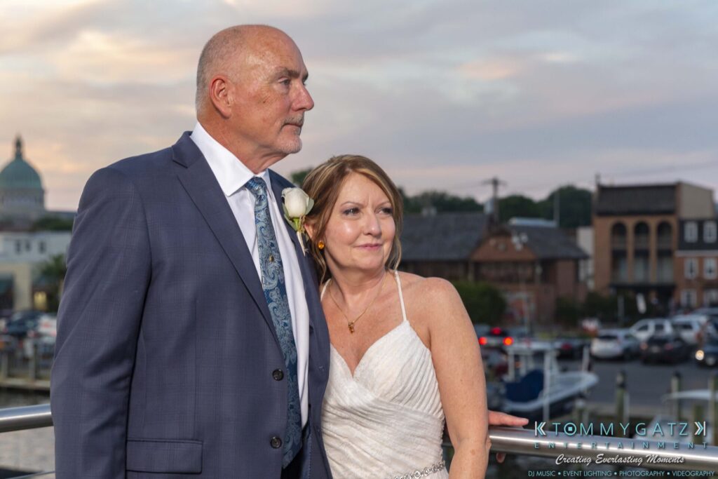 Grace-Michael-063018-RECEPTION-Low-Res-WM-508 - TOMMY GATZ ENTERTAINMENT A couple dressed in formal attire stands outdoors, with the man wearing a suit and the woman in a white dress. The background features buildings and a cloudy sky.