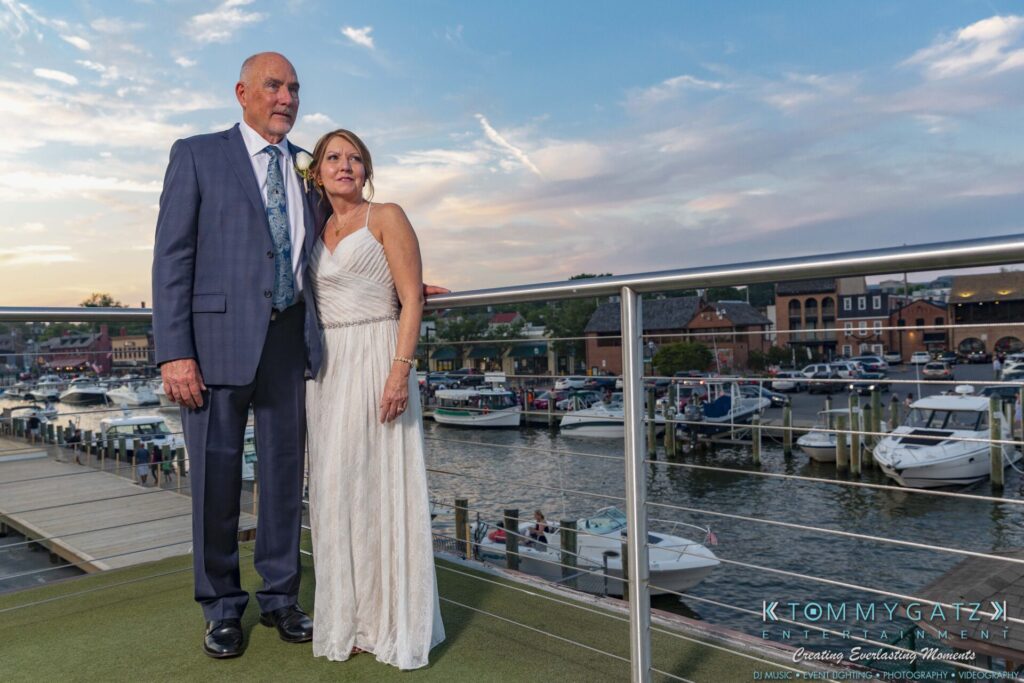Grace-Michael-063018-RECEPTION-Low-Res-WM-510 - TOMMY GATZ ENTERTAINMENT An elderly couple stands on a waterfront deck, dressed formally. The background features boats and a marina during sunset.