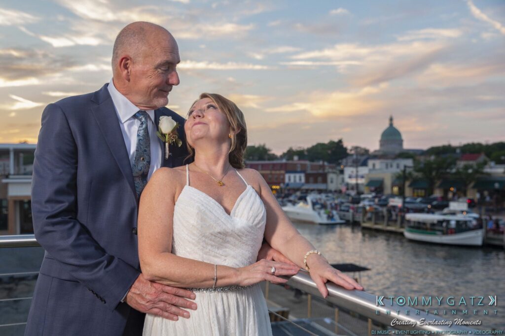 Grace-Michael-063018-RECEPTION-Low-Res-WM-511 - TOMMY GATZ ENTERTAINMENT A couple in wedding attire stand on a balcony overlooking a waterfront at sunset.
