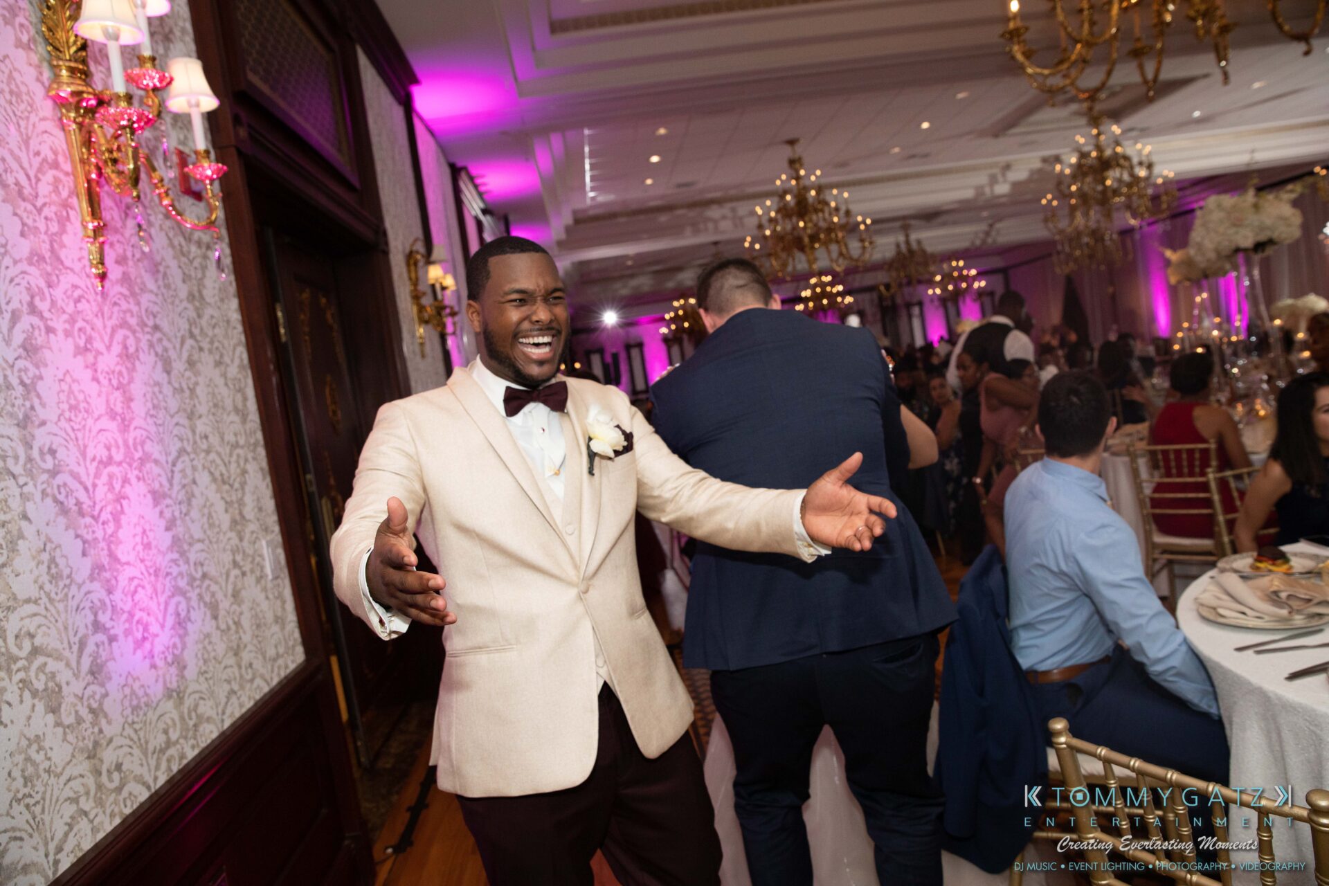 Harris-Wedding-HIGHLIGHTS-090119-101 - TOMMY GATZ ENTERTAINMENT A smiling man in a beige suit with a bow tie stands with open arms at an indoor event with chandeliers and decorated tables.