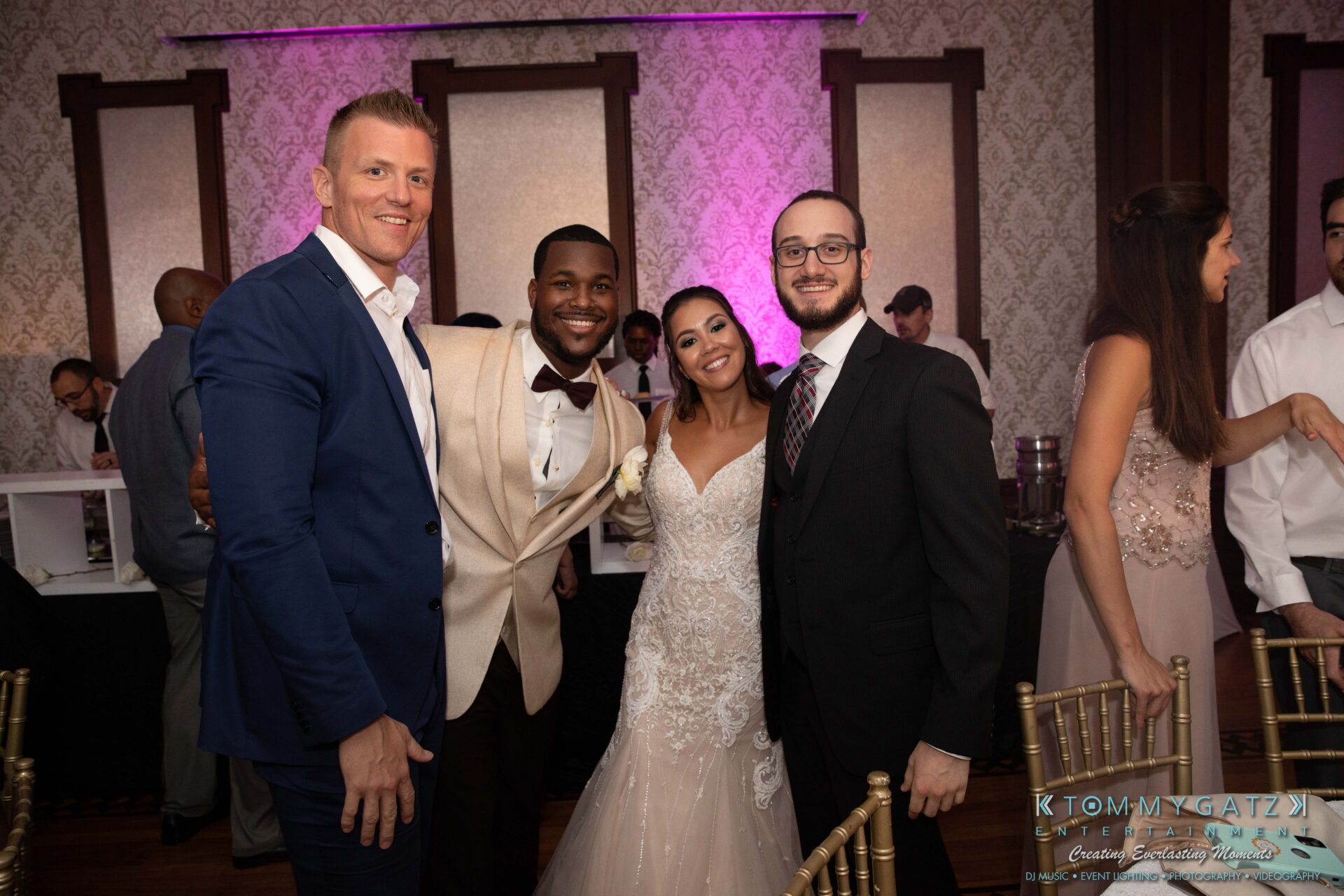 Harris-Wedding-HIGHLIGHTS-090119-103 - TOMMY GATZ ENTERTAINMENT Four people dressed in formal wear, including a bride in a white gown and a groom in a cream suit, stand smiling together in a warmly lit room with patterned walls.