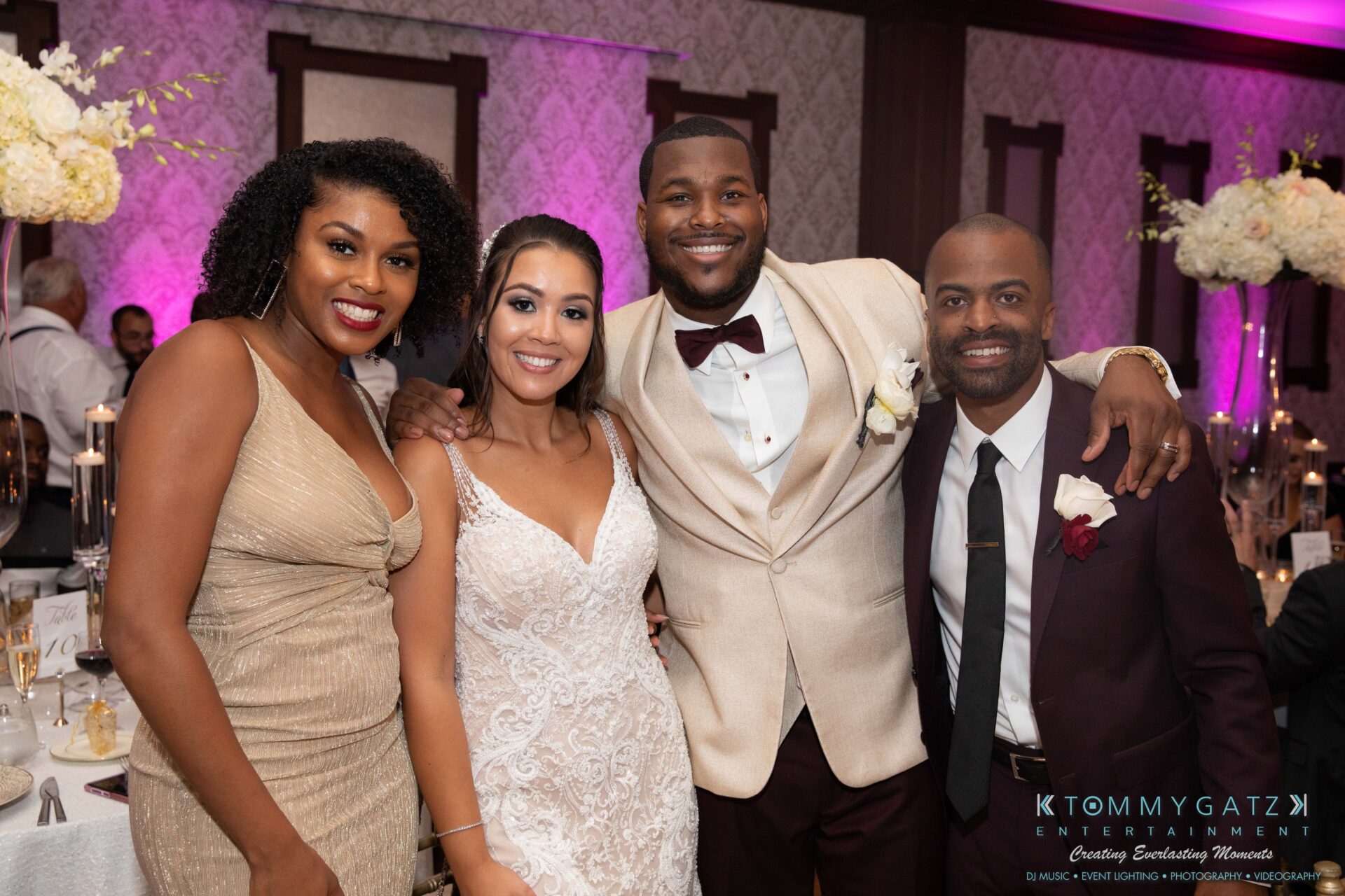 Harris-Wedding-HIGHLIGHTS-090119-104 - TOMMY GATZ ENTERTAINMENT Four people posing together at a formal event, with the two in the center dressed as a bride and groom. Dining tables and floral decorations are visible in the background.