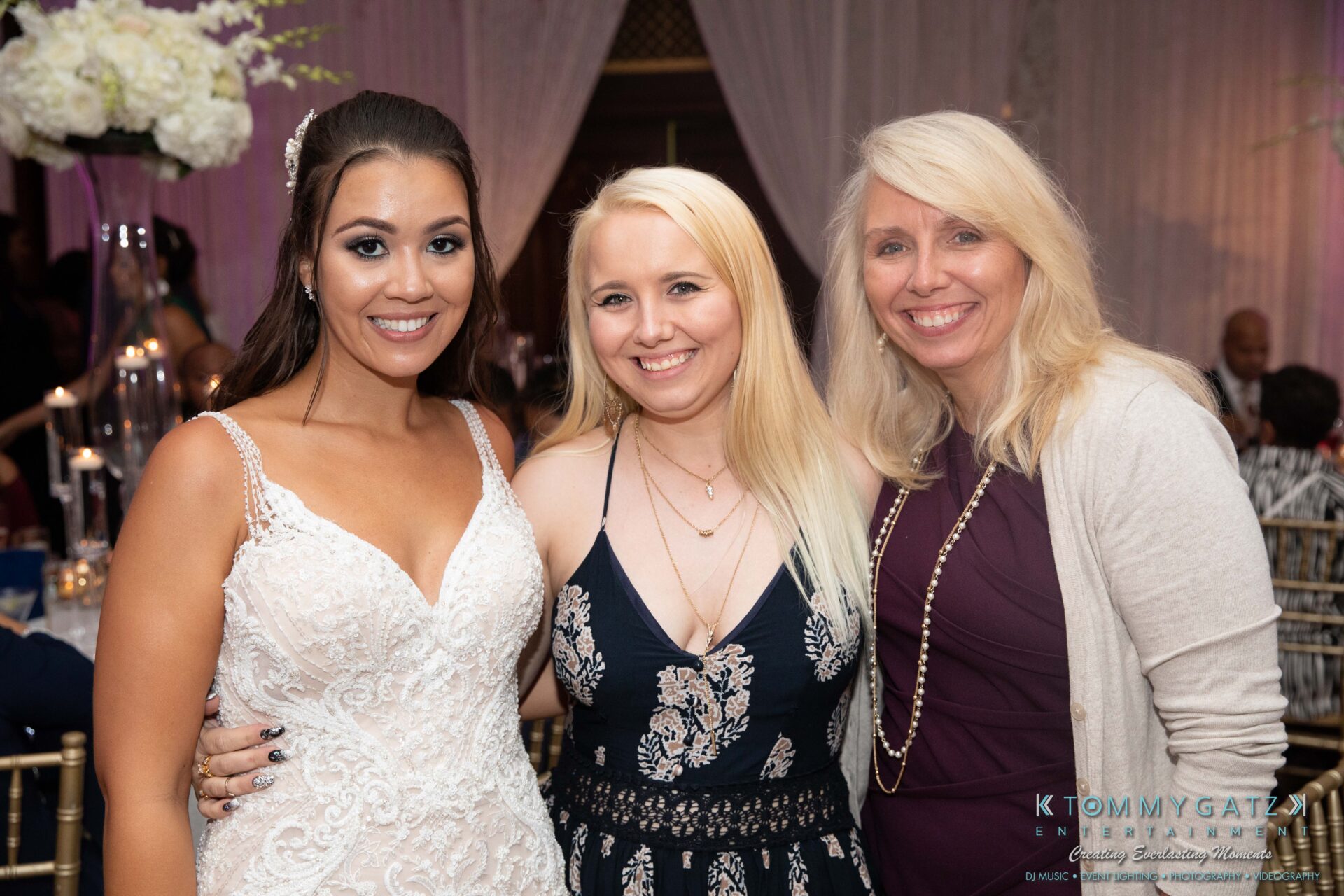 Harris-Wedding-HIGHLIGHTS-090119-108 - TOMMY GATZ ENTERTAINMENT Three women smiling and posing together at an indoor event, with decorated tables and guests in the background.