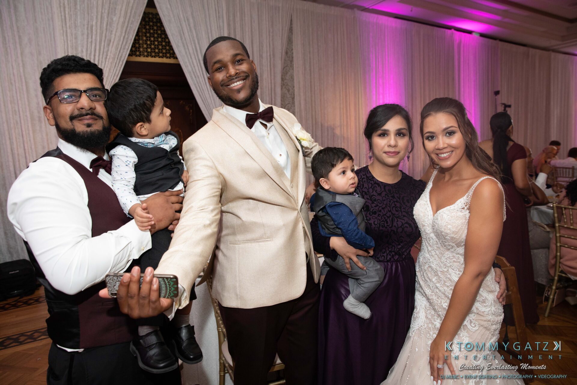 Harris-Wedding-HIGHLIGHTS-090119-110 - TOMMY GATZ ENTERTAINMENT Group of people dressed in formal attire at an event, with two adults holding young children. The background features decorative curtains and low lighting.