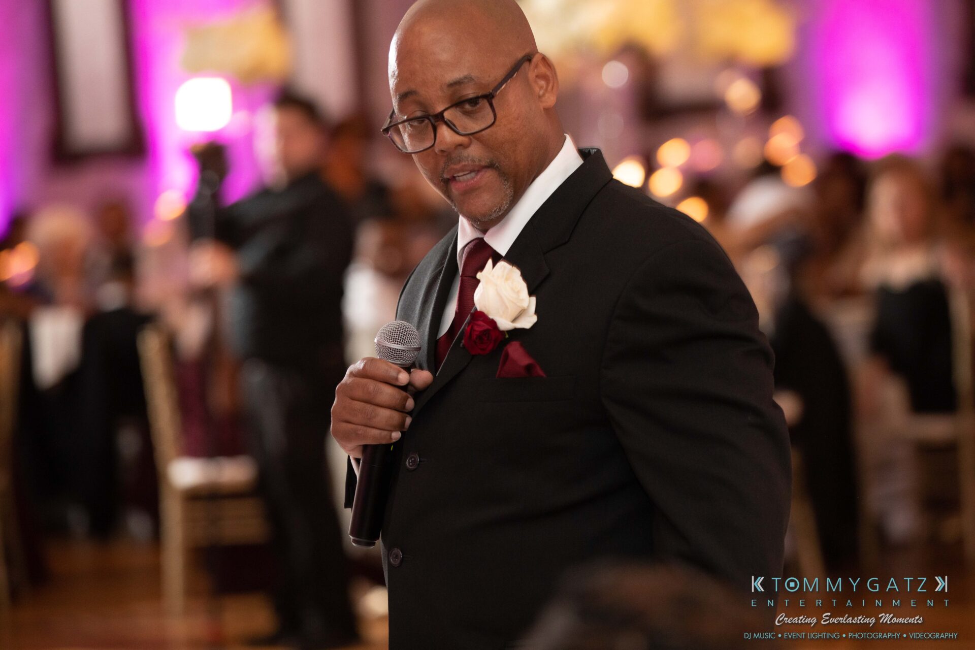 Harris-Wedding-HIGHLIGHTS-090119-117 - TOMMY GATZ ENTERTAINMENT A man in a suit holds a microphone at an indoor event with blurred people and pink lighting in the background.