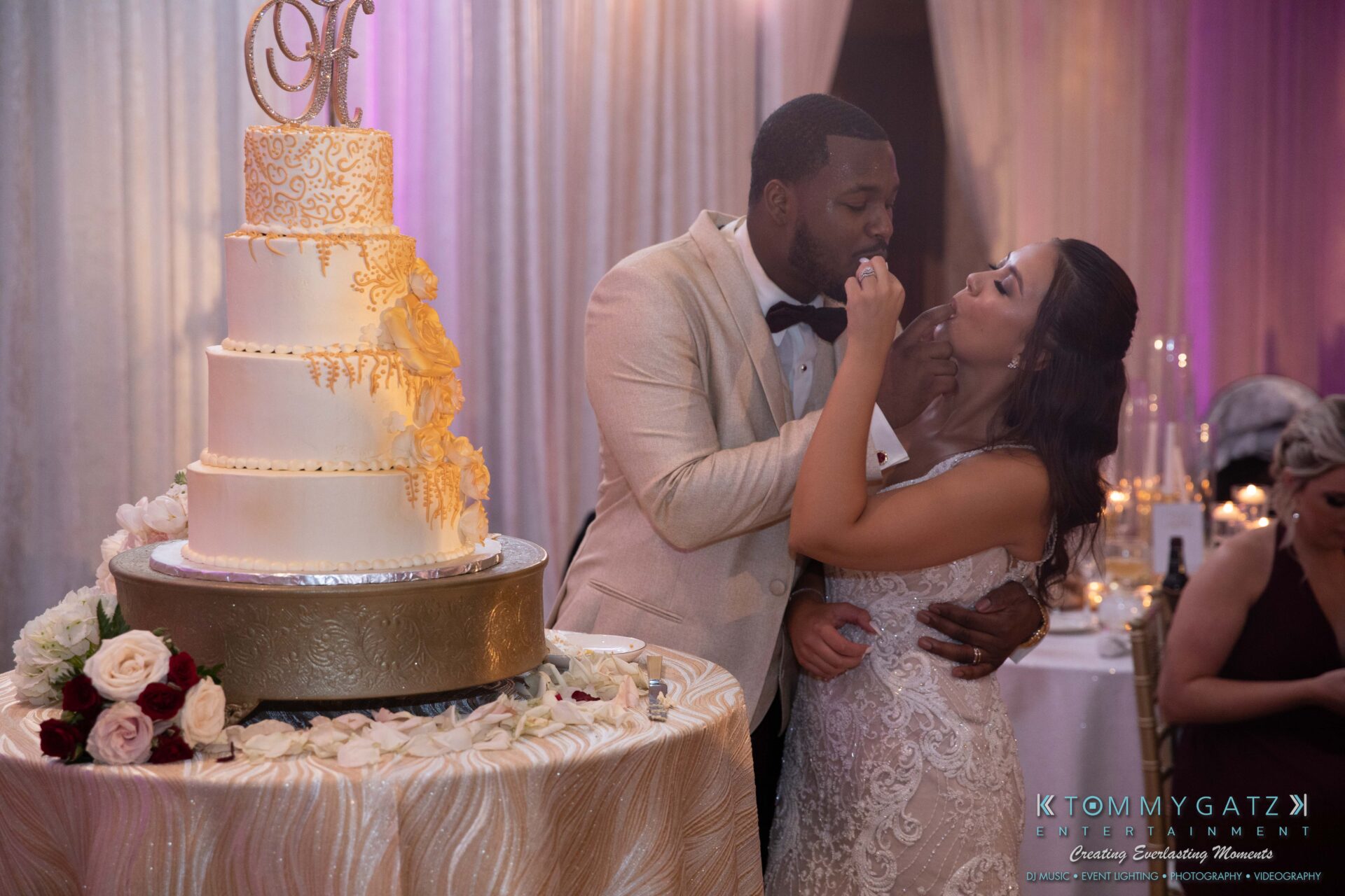 Harris-Wedding-HIGHLIGHTS-090119-123 - TOMMY GATZ ENTERTAINMENT A couple in formal attire shares a piece of cake at a wedding reception. A multi-tiered cake with gold accents is on the table.