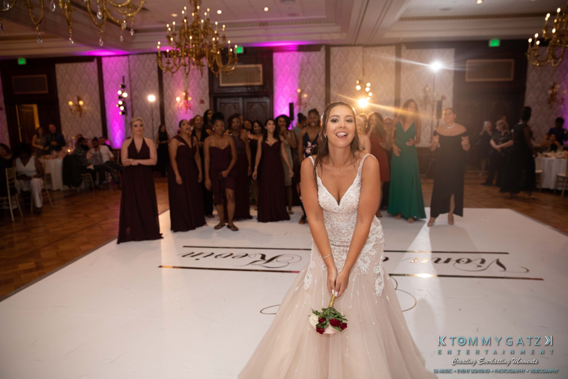 Harris-Wedding-HIGHLIGHTS-090119-125 - TOMMY GATZ ENTERTAINMENT A bride in a wedding dress prepares to throw a bouquet to a group of women on a dance floor. Chandeliers and guests are visible in the background.