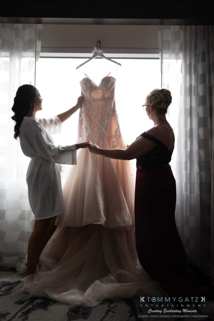 Harris-Wedding-HIGHLIGHTS-090119-13 - TOMMY GATZ ENTERTAINMENT Two women hold up a wedding dress in front of a window. The gown is lacey and illuminated by natural light.