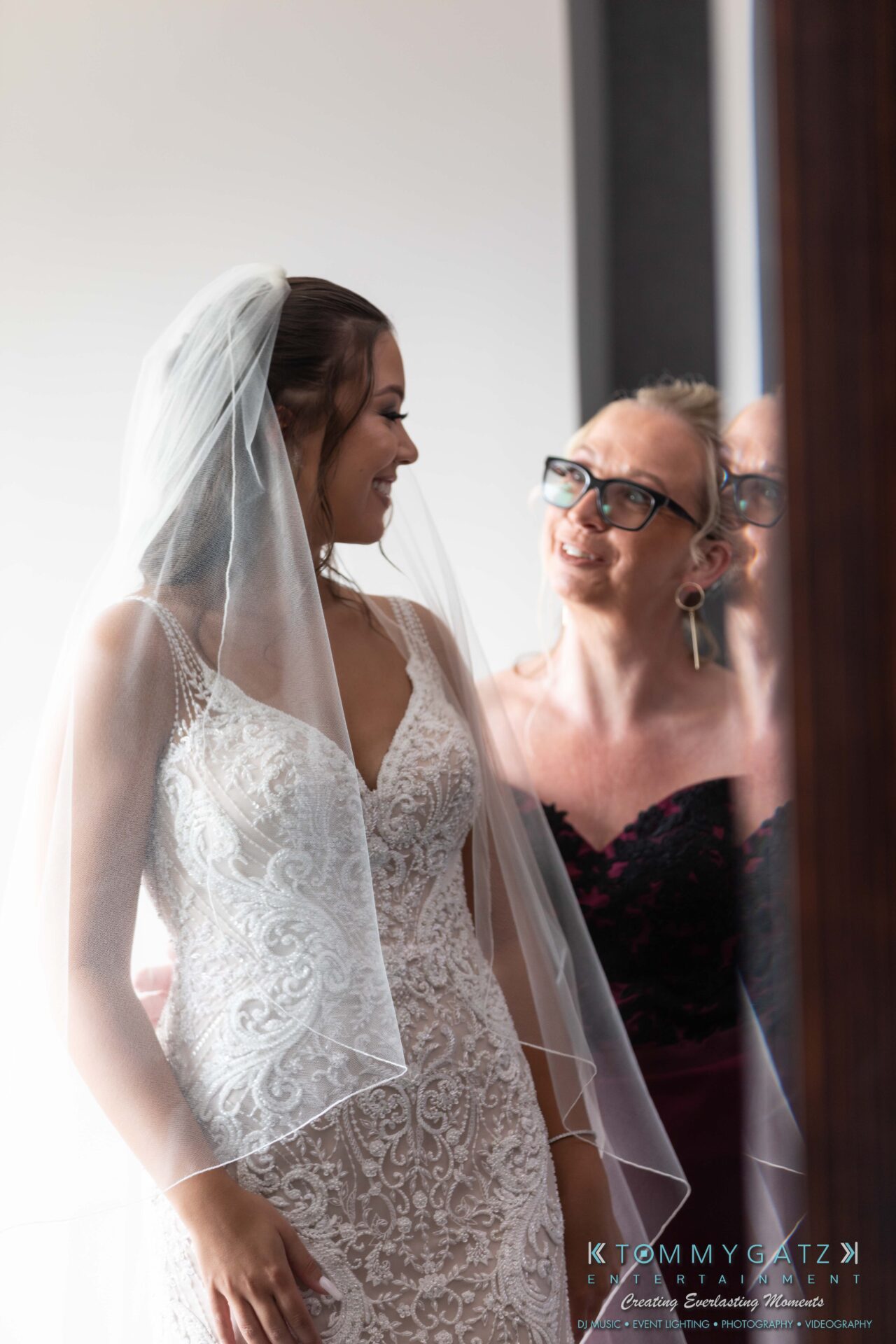 Harris-Wedding-HIGHLIGHTS-090119-28 - TOMMY GATZ ENTERTAINMENT Bride in a lace wedding dress and veil smiles at a person beside her, who is wearing glasses and a dark outfit. They are indoors, near a window with soft lighting.