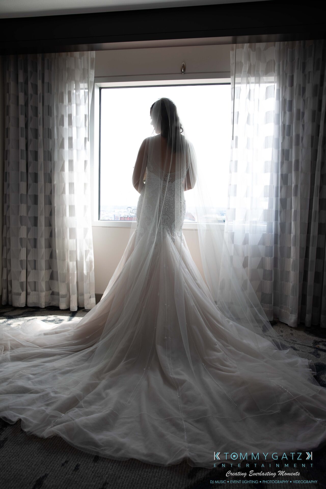 Harris-Wedding-HIGHLIGHTS-090119-32 - TOMMY GATZ ENTERTAINMENT A bride in a long, flowing gown stands facing a window with sheer curtains, allowing light to illuminate her silhouette.
