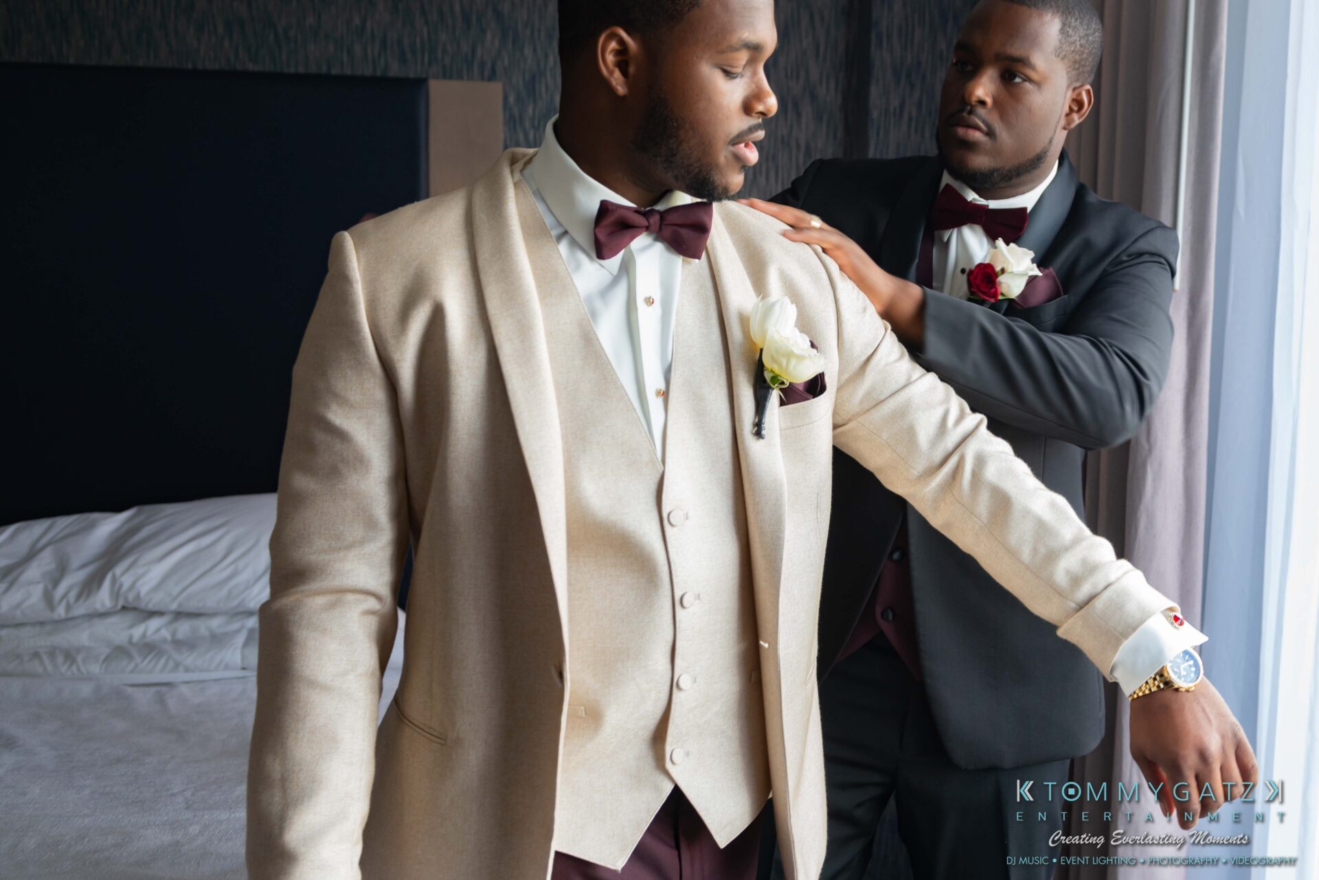 Harris-Wedding-HIGHLIGHTS-090119-45 - TOMMY GATZ ENTERTAINMENT Two men in formal attire; one is adjusting the other's beige jacket. They both wear bow ties and white floral boutonnieres, standing in a room with a bed in the background.