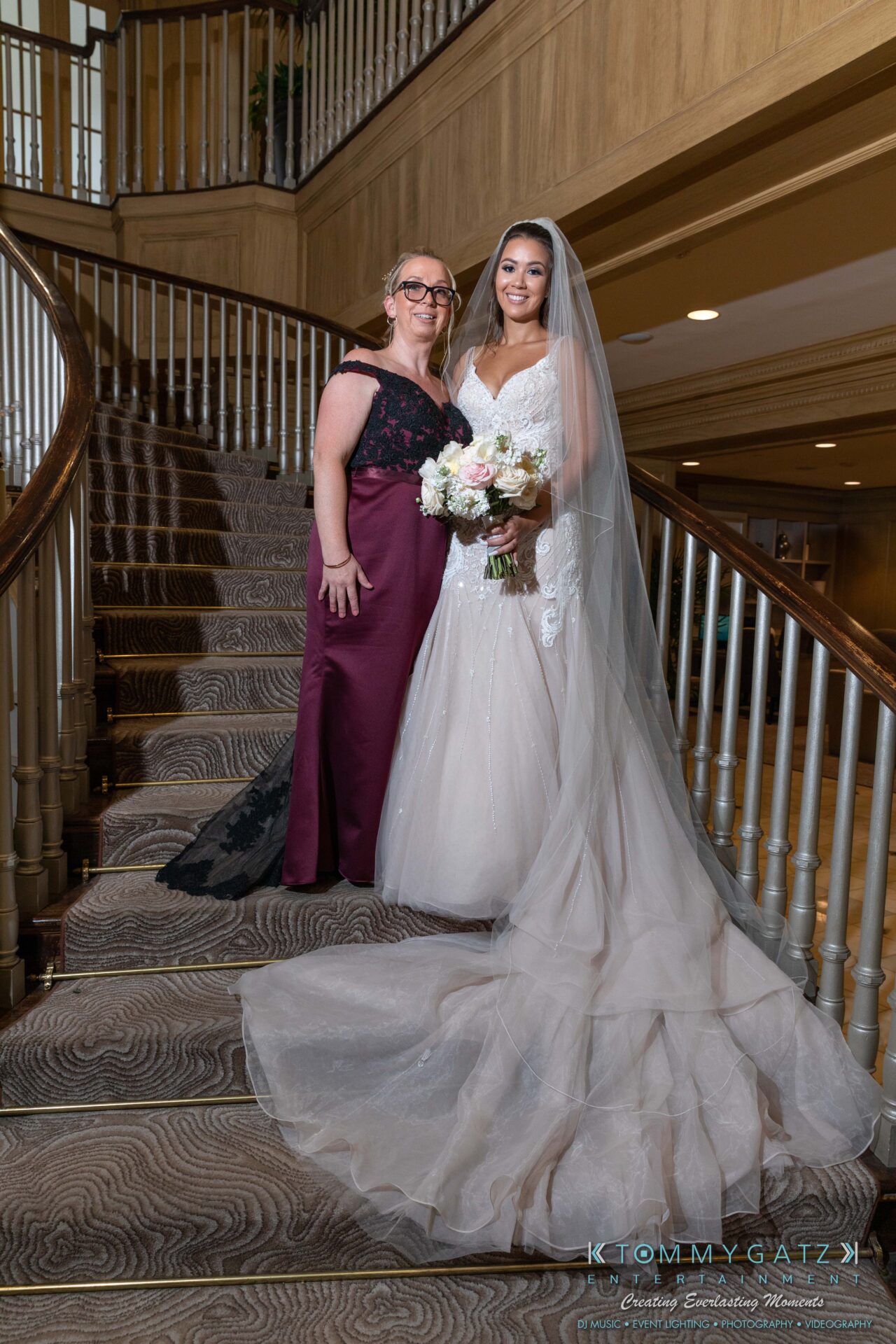 Harris-Wedding-HIGHLIGHTS-090119-47 - TOMMY GATZ ENTERTAINMENT Two women posing on a curved staircase indoors. One wears a wedding dress and holds a bouquet, while the other is in a formal gown.
