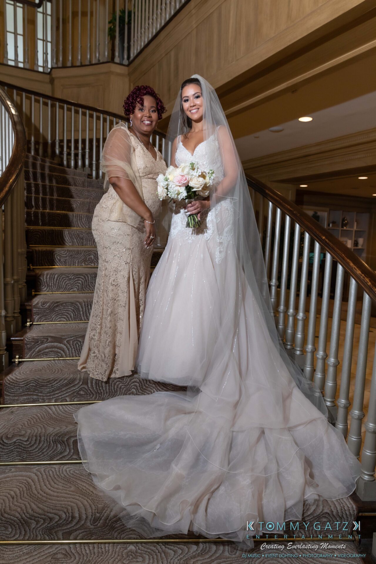 Harris-Wedding-HIGHLIGHTS-090119-48 - TOMMY GATZ ENTERTAINMENT Two women pose on a staircase; one is in a bridal gown holding a bouquet, the other is in an elegant dress.
