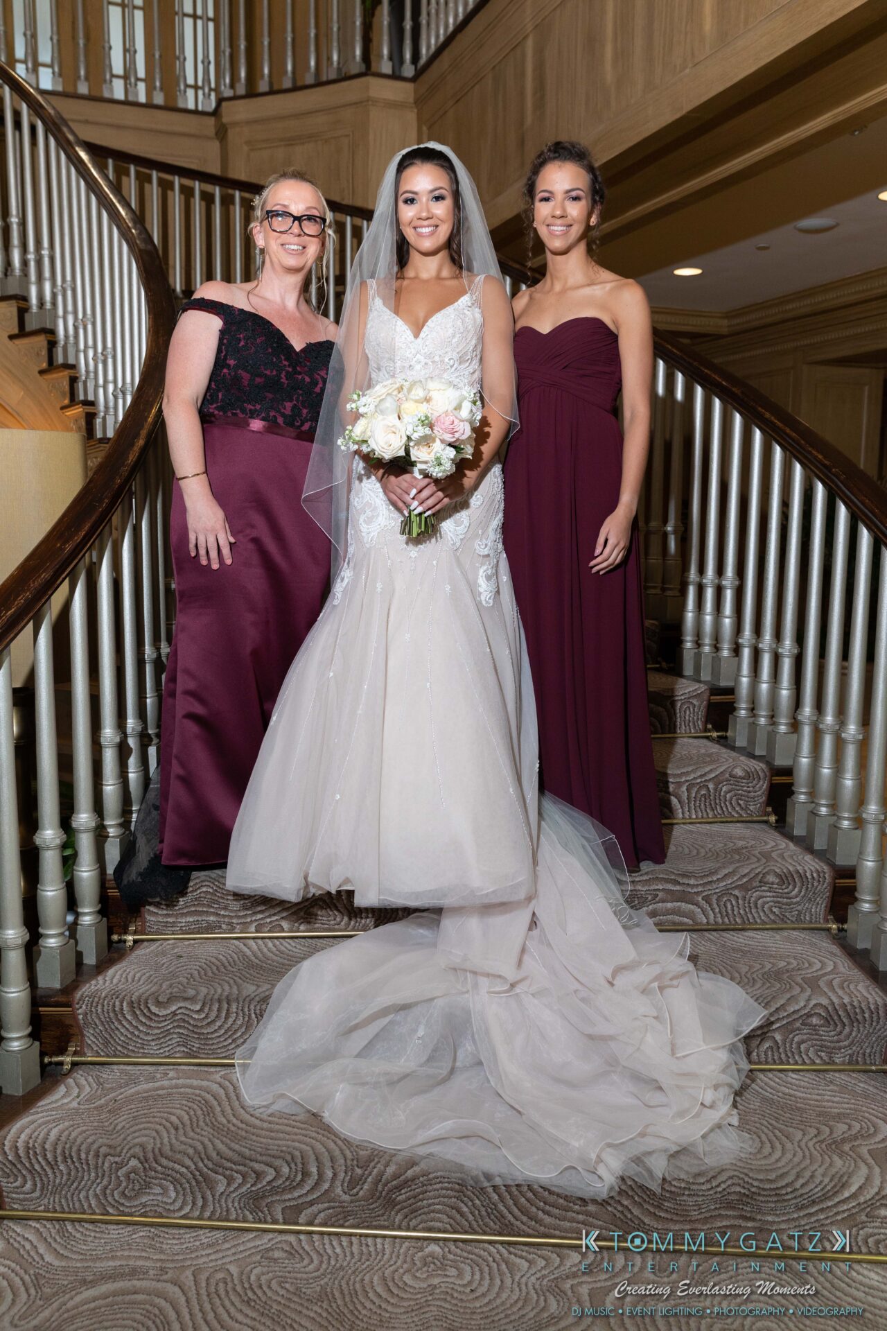 Harris-Wedding-HIGHLIGHTS-090119-50 - TOMMY GATZ ENTERTAINMENT Three women stand on a staircase, two wearing dark dresses and one in a white wedding gown holding a bouquet.