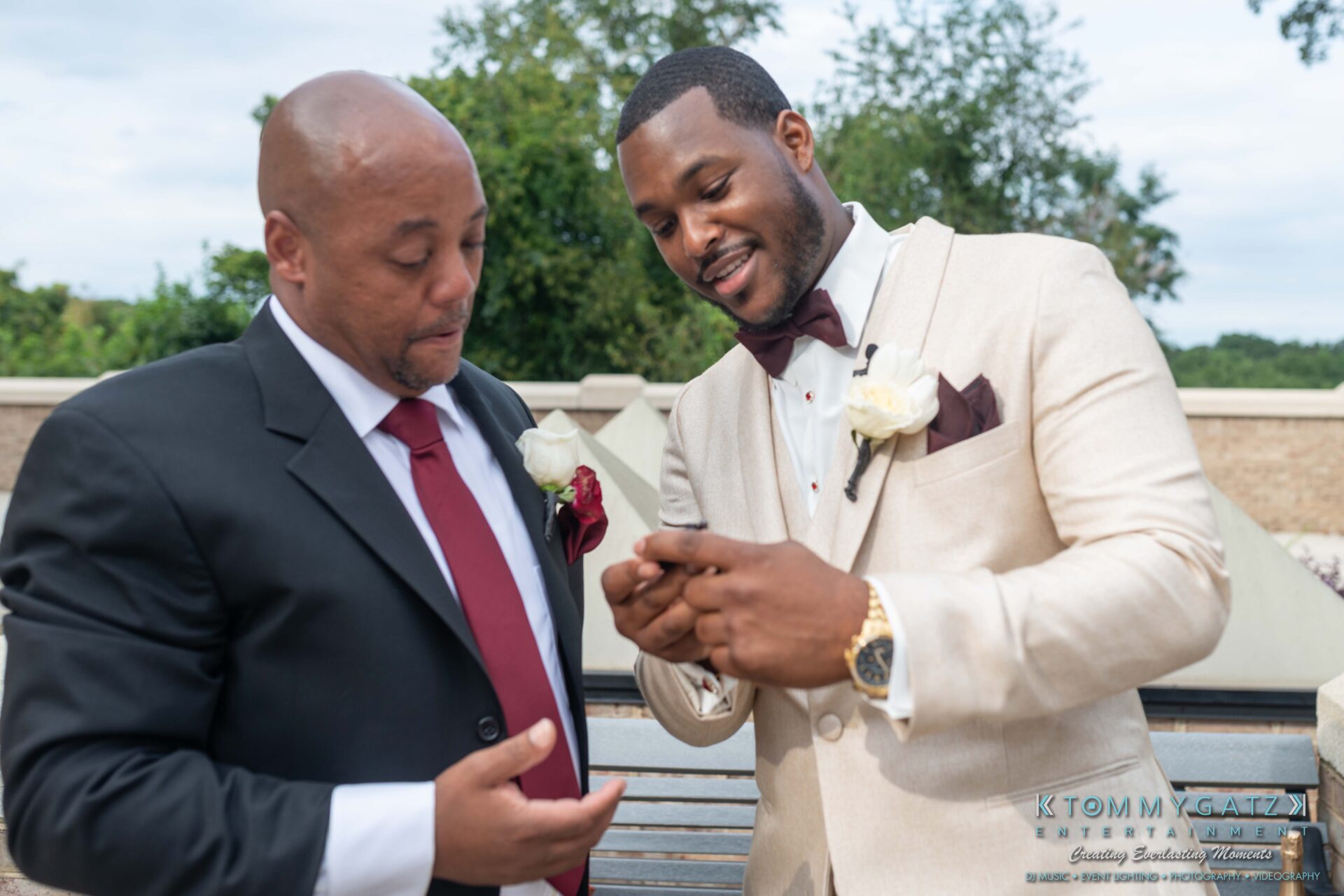 Harris-Wedding-HIGHLIGHTS-090119-57 - TOMMY GATZ ENTERTAINMENT Two men in suits stand outdoors looking at a smartphone. One wears a black suit with a red tie, the other a beige suit with a burgundy tie and boutonnière.
