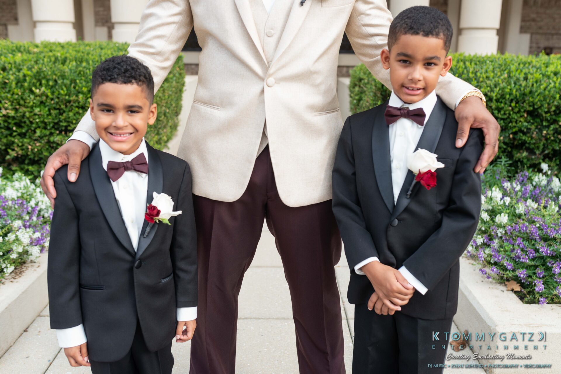 Harris-Wedding-HIGHLIGHTS-090119-58 - TOMMY GATZ ENTERTAINMENT Two young boys in suits with burgundy bow ties stand next to a man in a beige jacket and brown pants. They are outdoors, in front of a building with flowers in the background.