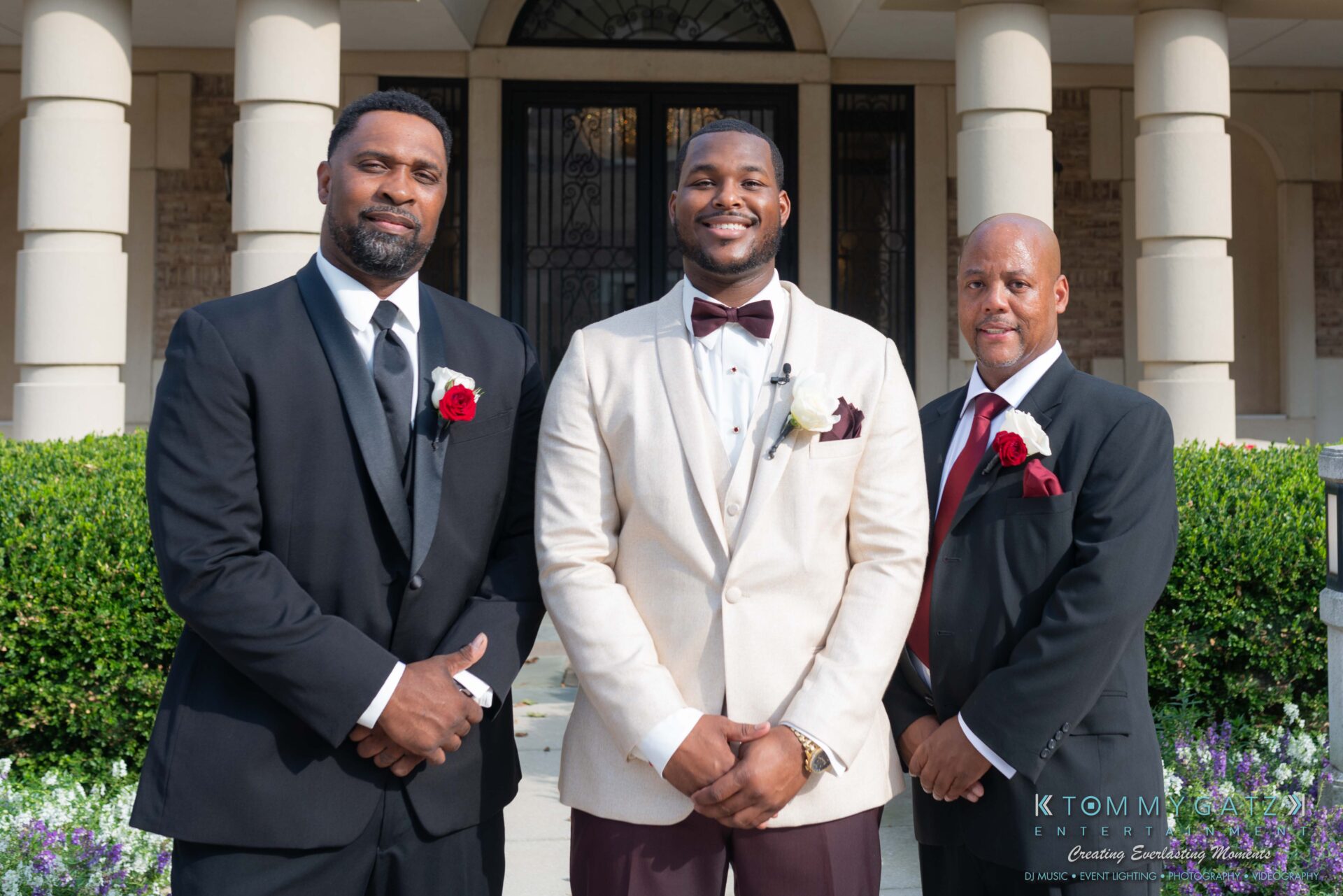 Harris-Wedding-HIGHLIGHTS-090119-60 - TOMMY GATZ ENTERTAINMENT Three men in formal attire stand in front of a building entrance. The man in the center wears a white suit, flanked by two men in black suits with red rose boutonnieres.