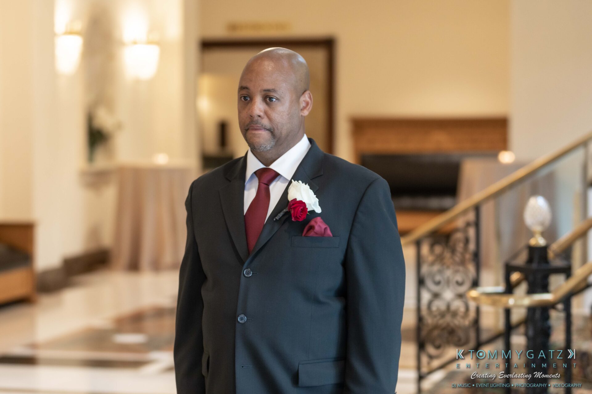 Harris-Wedding-HIGHLIGHTS-090119-65 - TOMMY GATZ ENTERTAINMENT A man in a dark suit with a white shirt and red tie stands in a hotel lobby, wearing a boutonniere.