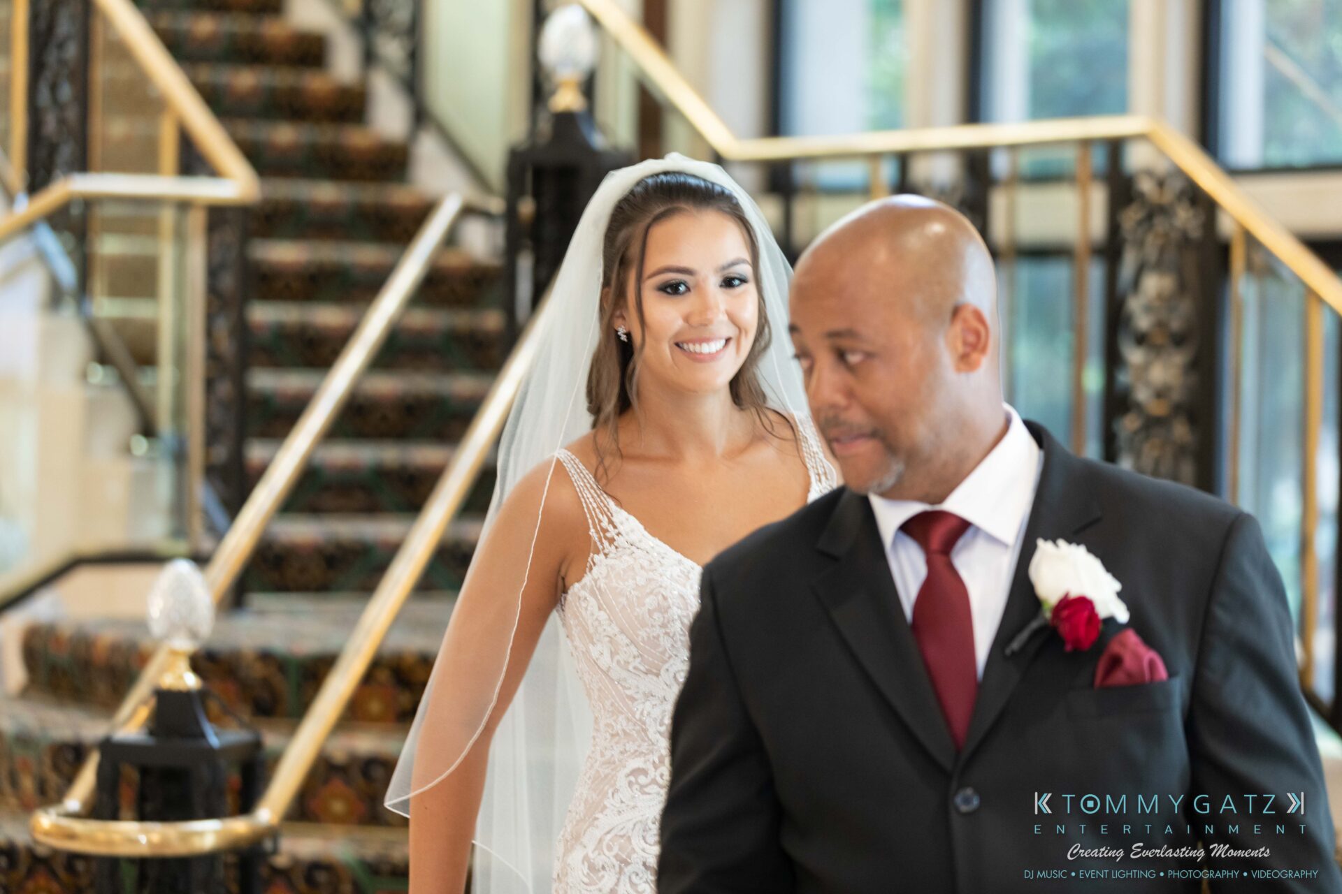 Harris-Wedding-HIGHLIGHTS-090119-66 - TOMMY GATZ ENTERTAINMENT Bride in a white dress smiles while standing behind a man in a suit with a red tie and boutonnière, set against a staircase background.
