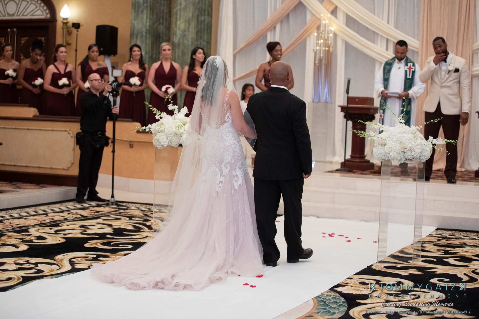 Harris-Wedding-HIGHLIGHTS-090119-70 - TOMMY GATZ ENTERTAINMENT A bride and her father stand before a wedding altar, facing the officiant. Bridesmaids in burgundy dresses stand to the left. White flower arrangements decorate the aisle.