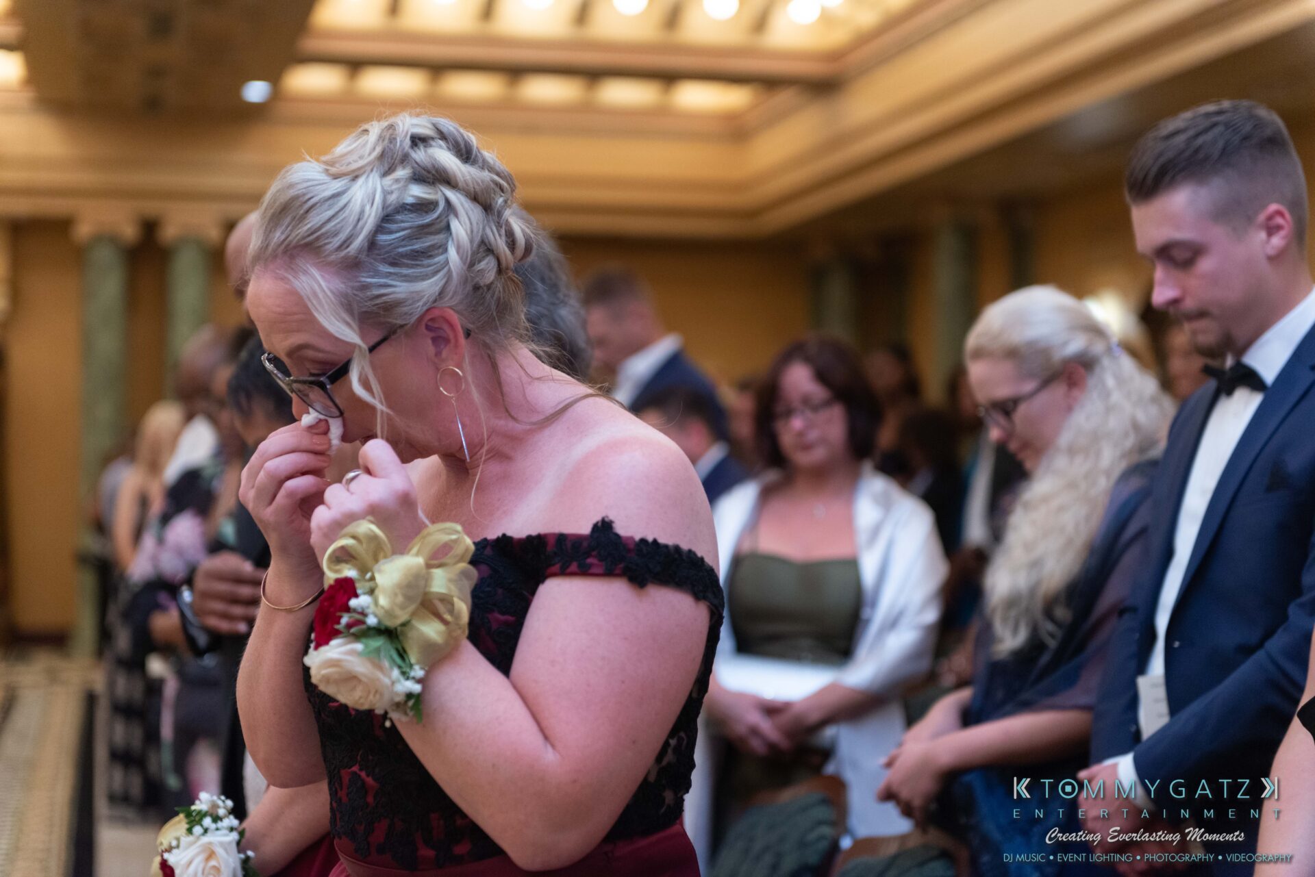 Harris-Wedding-HIGHLIGHTS-090119-74 - TOMMY GATZ ENTERTAINMENT A woman in a formal dress wipes tears during a ceremony, with people seated and standing in the background.