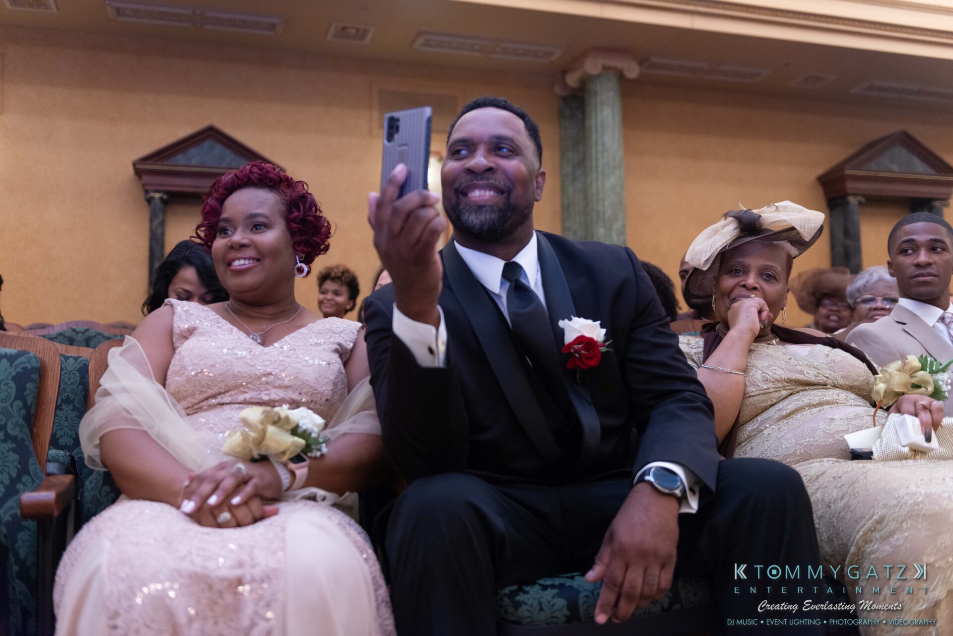 Harris-Wedding-HIGHLIGHTS-090119-75 - TOMMY GATZ ENTERTAINMENT A group of people in formal attire sit in a row. A man in a suit holds a smartphone up, smiling. Others appear attentive and smiling.