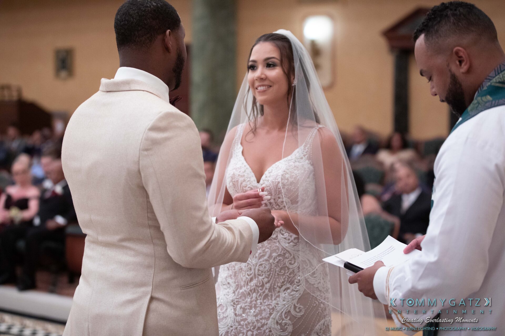 Harris-Wedding-HIGHLIGHTS-090119-77 - TOMMY GATZ ENTERTAINMENT A bride and groom exchange rings during a wedding ceremony, both dressed in formal attire. The bride wears a veil and lace gown, while the groom is in a light-colored suit.