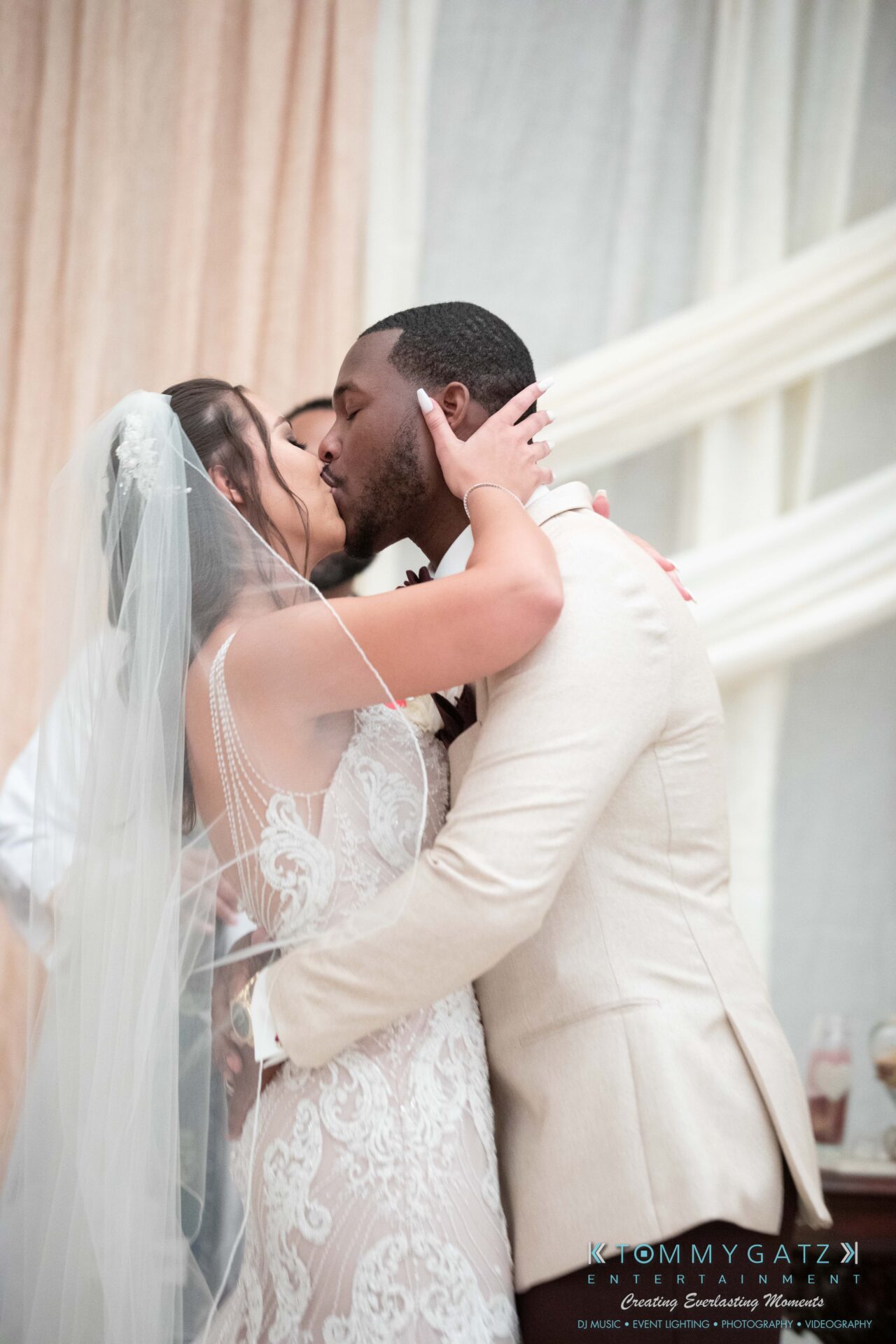 Harris-Wedding-HIGHLIGHTS-090119-83 - TOMMY GATZ ENTERTAINMENT Bride and groom share a kiss wearing wedding attire. Bride in lace dress and veil, groom in beige suit.
