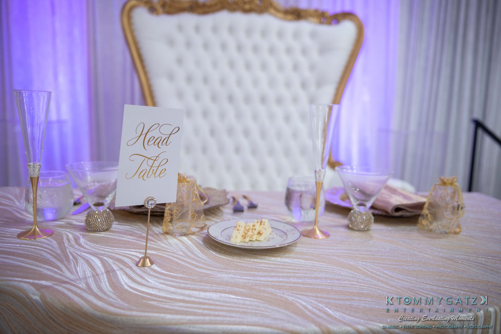 Harris-Wedding-HIGHLIGHTS-090119-86 - TOMMY GATZ ENTERTAINMENT Elegant head table setup with a gold-accented chair, champagne glasses, a plate with cake slices, and a sign reading "Head Table" on a shimmering tablecloth.